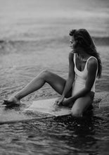 Load image into Gallery viewer, A stunning black and white photograph titled &#39;Riding Tide,&#39; showcasing a woman in a white swimsuit sitting on a surfboard at Wilmington Beach, North Carolina, capturing the essence of serene ocean vibes.
